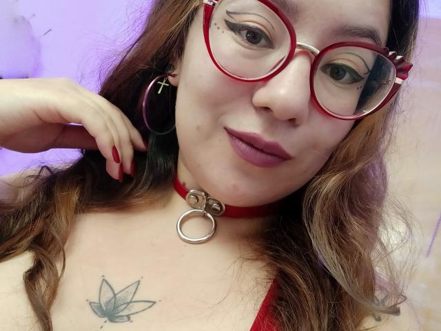 HollyManziniBBW with red glasses, red choker, ring, and hoop earrings, red lipstick, tattoo on chest, wavy brown hair, purple eyeliner, and red nails, smiling.