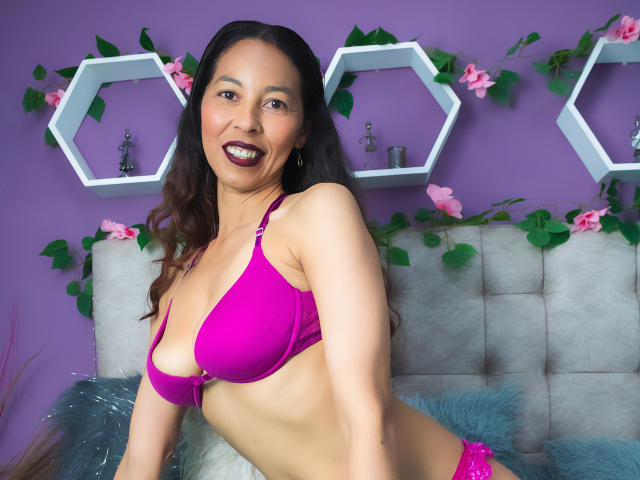 RosalindaCortez with dark brown hair, purple bra, and matching panties, smiling at the camera, showing her small, perky tits, and flat, sexy ass, against a purple wall.