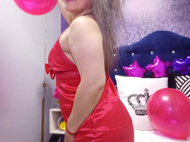 BiancaWild mit rosa Ballons, trägt ein rotes Satin-Dessous-Kleid, zeigt ihr Gesäß und ihre Brüste, langes braunes Haar, lächelnd, steht in einem Schlafzimmer mit einem blauen Kopfteil..