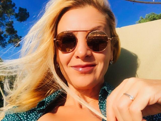 Aliciya Sage, met blond haar, draagt ronde aviatorzonnebrillen, een groene top met een patroon, en een diamantenring. Ze glimlacht met glanzende lippen, met zonlicht in haar haar, tegen een blauwe lucht..