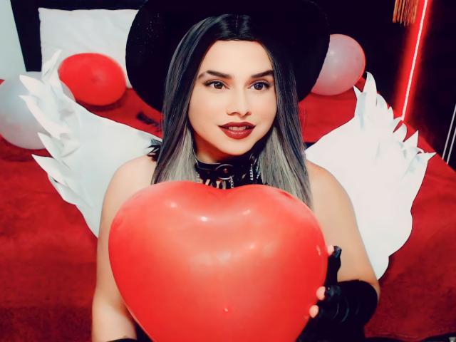 CamilaFetishDirty with gray hair, black hat, and white angel wings, holding a red heart, wearing a black collar, and black gloves, with a sexy, confident expression.