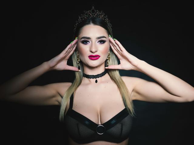 LoraDevine, con i capelli biondi, indossa un reggiseno di rete nero con anello metallico, un collare nero e una tiara, con le mani sulle orecchie, rossetto rosa, unghie verdi, sfondo scuro..