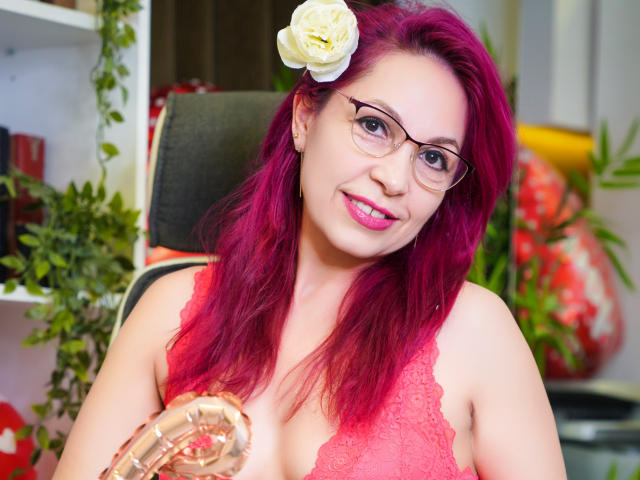 DirtyMature with pink hair, white flower in her hair, glasses, red lace top, holding a shiny balloon, smiling at the camera. Boobs visible, pink lipstick, cute.