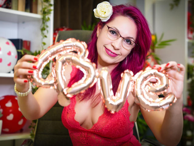 DirtyMature with pink hair, glasses, and flower in her hair, holding "love" balloons, wearing a red lace top, smiling, showing her tits, and red nails.