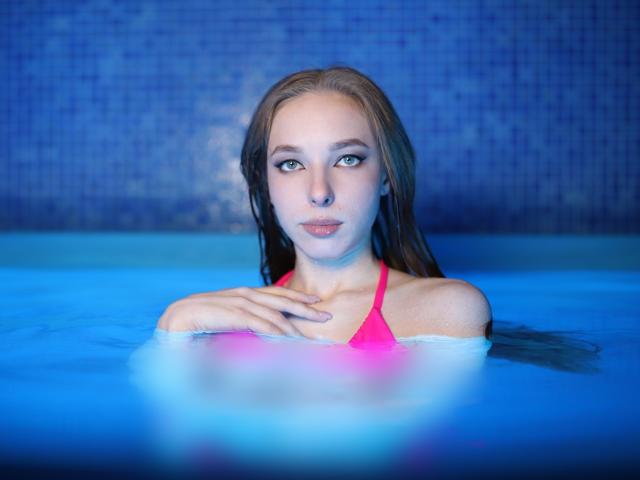 El top de bikini rosa y mojado de SafiraFuracao, sus pequeños senos y su piel pálida son visibles en una piscina con azulejos azules, con su mano sobre el pecho y una expresión tranquila y seria..