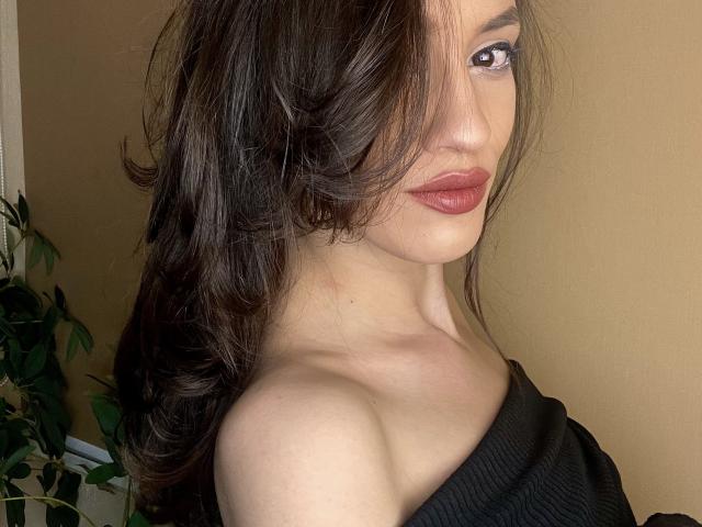 VievienneClemente with dark wavy hair, brown eyes, and red lipstick, wearing an off-shoulder black top, showing bare shoulders. Her face is slightly turned, looking at the camera.