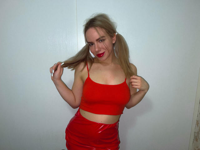 CandyWithBrandy met vlechtjes, die een rode crop top en een glimmende rode rok draagt, haar haar vasthoudend, met rode lippenstift, waarbij haar borsten en billen zichtbaar zijn..