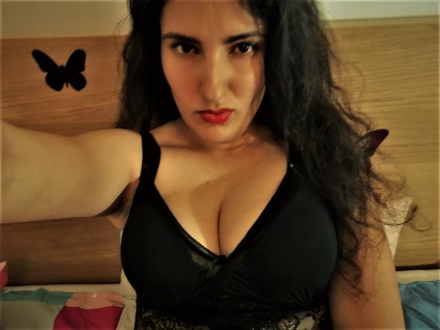 AddictedToUTS with curly black hair, red lipstick, and black bra showing big boobs, taking a selfie, wood headboard in background, black butterfly sticker on left.