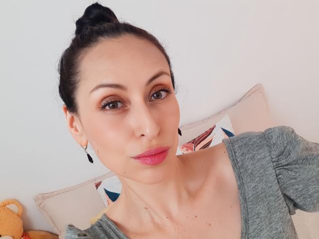 AliceRossi has a cute face with pink lips, brown eyes, and black hair in a bun. She's wearing a grey top and black earrings, looking at the camera.