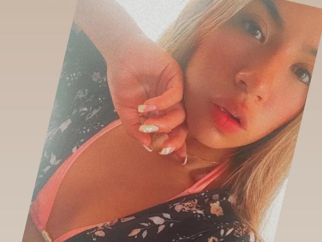 ChloeDeluxee, con i capelli biondi, indossa lingerie rosa, un abito nero con fiori, tiene il mento, ha labbra rosa lucide, unghie decorate e un'espressione carina, leggermente seducente. Il suo seno è visibile in modo discreto..