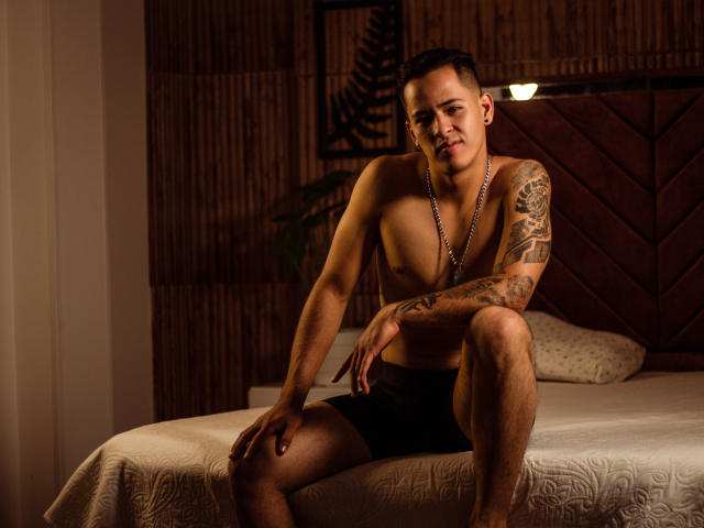 AbbyDrake is een gespierde, getatoeëerde man met kort donker haar, die zwarte boxershorts draagt en op een bed zit. Hij kijkt met een lichte glimlach in de camera en draagt een ketting..