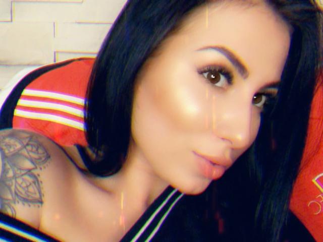 MindyDesire tiene el pelo negro, ojos marrones y un tatuaje en el hombro izquierdo. Lleva una camiseta negra con un fondo a rayas rojas y blancas..
