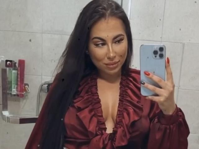MindyDesire, avec ses longs cheveux noirs, portant un haut rouge vif à volants, montrant sa poitrine dans un selfie pris devant un miroir, tenant un téléphone argenté avec des ongles rouges..