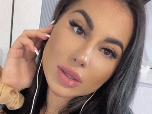 MindyDesire tiene labios rosados y brillantes, cejas oscuras, pestañas largas y lleva auriculares blancos, con la mano cerca de la oreja, mirando ligeramente hacia arriba. Tiene un tono de piel bronceado..