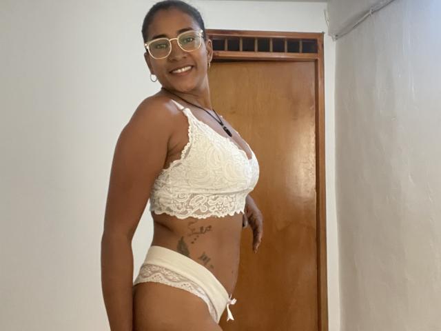 Thannia with dark skin, glasses, white lace bra, and panties, showing her tits and pussy tattoos, smiling, standing in a room.