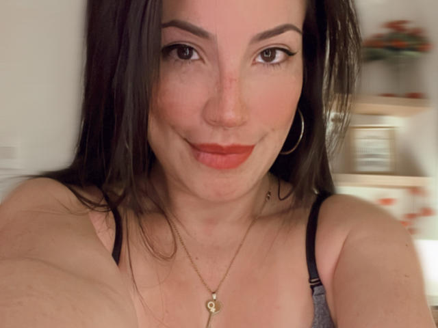Liliysweet a les cheveux brun foncé, du rouge à lèvres, et elle sourit à l'appareil photo. Elle porte un soutien-gorge noir avec des boucles d'oreilles dorées et un collier. Ses seins sont visibles..