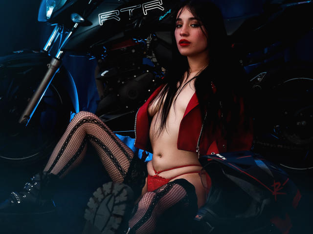 DulceHarper, com cabelos longos e pretos, jaqueta vermelha cropped, calcinha de renda vermelha, meia-calça de rede, botas pretas, sentada em uma motocicleta, expondo seus seios e vulva, em um ambiente escuro e melancólico..