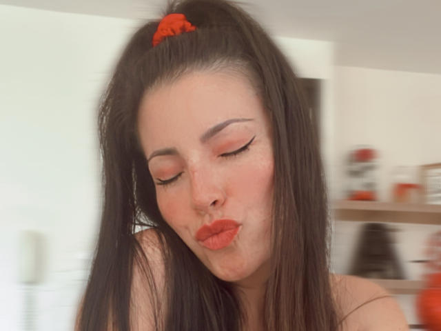 Liliysweet has long black hair in a red scrunchie, closed eyes, and red lipstick, puckering her lips. She has fair skin, pink eyeshadow, and subtle freckles.