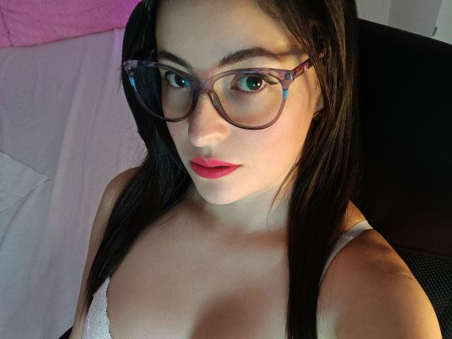 VaaleryStark with glasses, red lips, and straight black hair, showing her tits in a white lace top, sitting in a black chair.