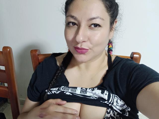 SakotoDika with brown skin, braided hair, pink lipstick, black shirt, lifting shirt to expose her tits, sitting on wooden chair, wearing colorful earrings, smiling slightly.