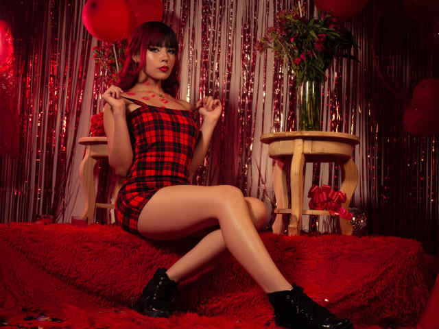 MagicianFoster, con un vestito a quadri rossi, stivali neri, che tiene una frusta, ha il rossetto rosso, i capelli rosso scuro, ed è seduta su una superficie rossa e pelosa, mostrando i suoi seni e il suo sedere..