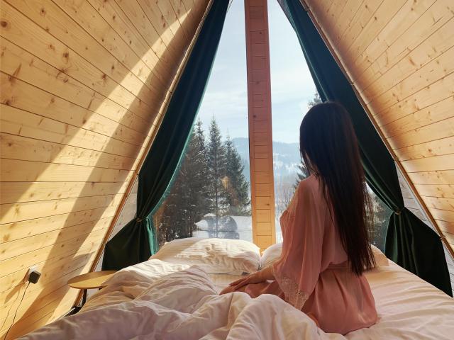 LauraineX with long brown hair sits on a bed, wearing a pink robe, facing a window with green curtains, showing her back and ass. Wooden walls, white bedding.