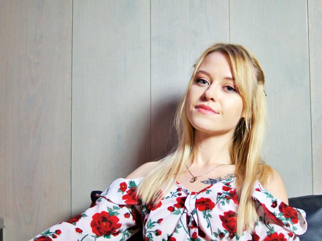 CindyxGlam mit blonden Haaren, trägt ein blumiges Oberteil mit einem Schulterauschnitt, hat kleine Brüste und eine Halskette, lächelt leicht und sitzt an einer Holzwand..