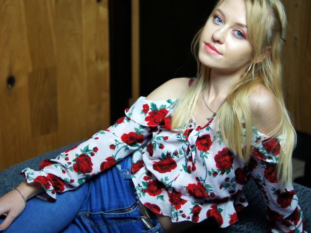 CindyxGlam mit blonden Haaren, blauen Augen, trägt ein rotes, blumiges Oberteil mit offener Schulter und blaue Jeans, lehnt sich leicht zurück und lächelt entspannt und verführerisch. Die Brüste sind teilweise sichtbar..