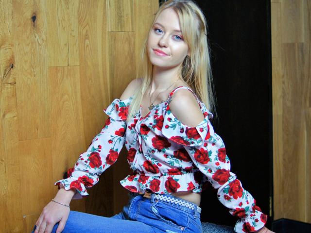CindyxGlam has blonde hair, blue eyes, and a cute smile, wearing a floral off-shoulder top and blue jeans, sitting with a slight tilt, showing her small tits.