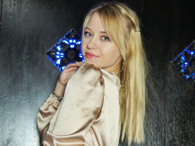 CindyxGlam with blonde hair, blue eyes, and pink lipstick, wearing a satin beige top, smiling at the camera, showing her ass with her hand near her face.