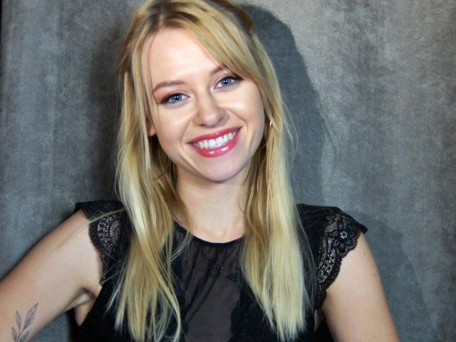 CindyxGlam has blonde hair, blue eyes, and is smiling with red lipstick. She's wearing a black lace top, showing a small tattoo on her left arm.