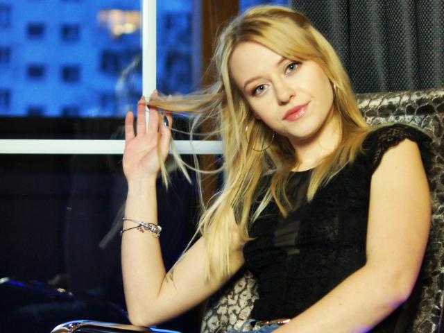CindyxGlam with blonde hair, black lace top, and hoop earrings, holding a strand of her hair, smiling, wearing a bracelet, seated indoors, blue city lights in background.