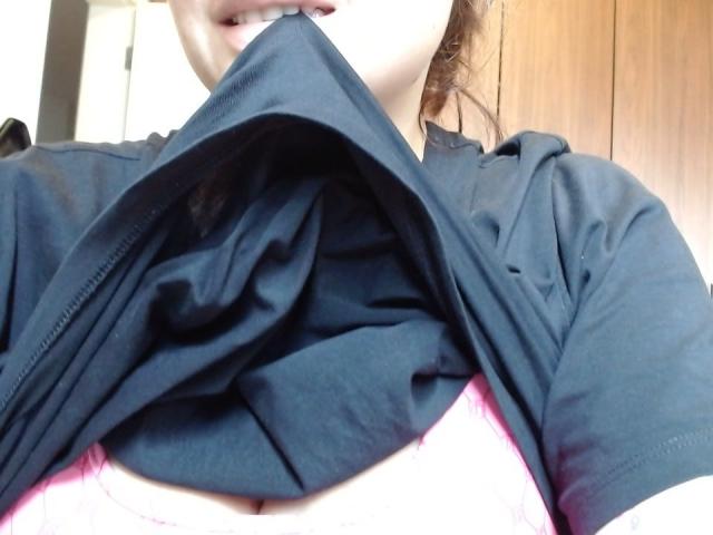 LuxEsa soulève un t-shirt noir pour montrer des bonnets de soutien-gorge roses, exposant ses seins. Sa bouche est légèrement ouverte et ses dents sont visibles. Son visage est partiellement visible..