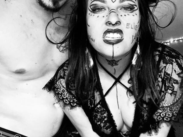 Schwarz-weißes Foto von DagmarEtMirana mit Gothic-Make-up, blutigen Lippen, einem Spitzenoberteil und freiliegenden Brüsten, neben einem Mann ohne Hemd. Sie blickt mit einem Knurren nach oben..