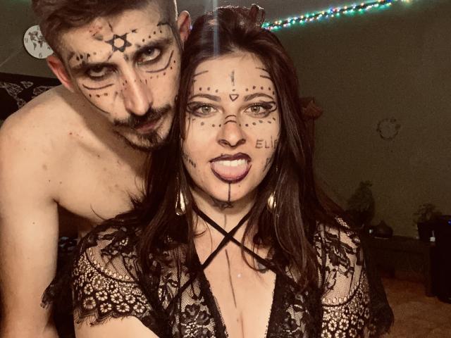 DagmarEtMirana e un uomo con il viso dipinto di nero, tatuaggi e barba, in piedi dietro di lei, senza camicia. Lei indossa pizzo nero, ha il seno scoperto e una collana nera a forma di croce..