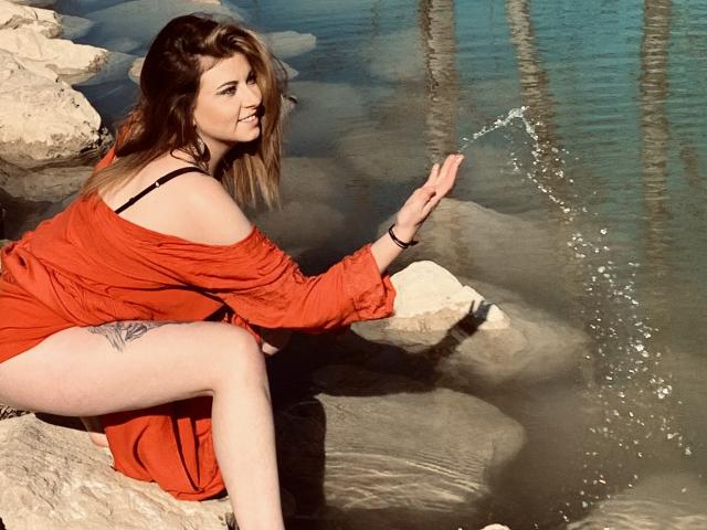 DagmarEtMirana with brown hair, wearing an orange dress, black strap showing, sitting on rocks, splashing water with her hand, visible tattoo on right thigh, smiling.