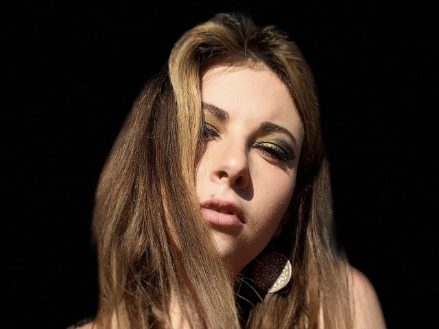 DagmarEtMirana tiene el cabello rubio oscuro, sombra de ojos verde y la boca ligeramente abierta. Su nariz está centrada y lleva grandes aretes redondos sobre un fondo oscuro..