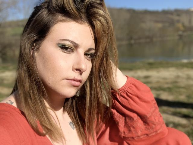 DagmarEtMirana avec des cheveux bruns, du fard à paupières vert, une robe rouge, un piercing au nez et un tatouage, regardant vers le bas, la main dans les cheveux, la lumière du soleil sur le visage, en extérieur, arrière-plan légèrement flou..