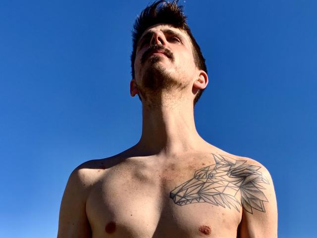 DagmarEtMirana, ohne Hemd, mit einem Wolfstattoo auf der rechten Schulter, blickt nach oben, hat kurze, dunkle Haare, einen leichten Bart und blasse Haut vor einem klaren, blauen Himmel..