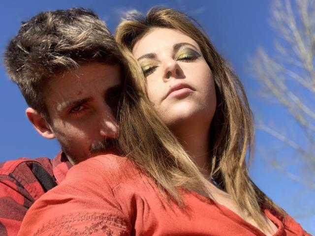 DagmarEtMirana met groene oogmake-up, bruin haar en een rood shirt, staan voor een man met een baard en kort bruin haar, die een rood geruit shirt draagt..