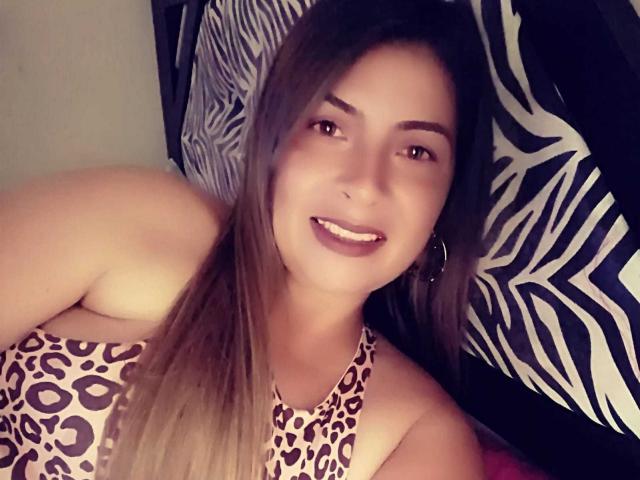 BrendaJess, com cabelos castanhos longos, olhos castanhos e brincos de argola, sorrindo, vestindo uma blusa de alça com estampa de leopardo, mostrando seus ombros e parte superior do peito, deitada em um travesseiro com padrão de zebra..