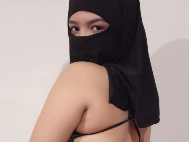 Maryame, avec un masque noir recouvrant sa bouche et ses yeux, portant un foulard noir, un soutien-gorge à bretelles noires, laissant apparaître son épaule nue et le haut de son dos, regardant par-dessus son épaule..