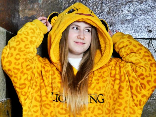 MeganCandy with long blonde hair, wearing a yellow leopard-print hoodie with an animal head, hands in hood, smiling, against a textured wall.