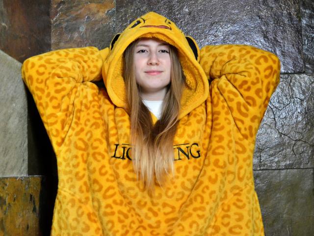MeganCandy indossa una felpa gialla con stampa leopardo e cappuccio a forma di Pikachu, con le braccia alzate, lunghi capelli biondi, un leggero sorriso, in piedi di fronte a un muro con una superficie strutturata..
