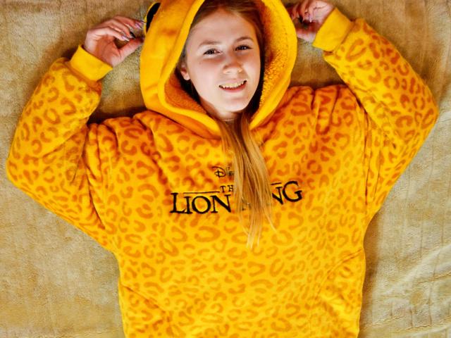 MeganCandy with blonde hair, smiling, wearing a yellow leopard print hoodie with a hood up, arms raised, lying on a beige textured surface. Her tits are small.