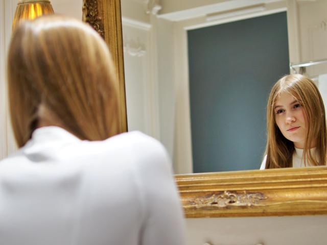 MeganCandy, con lunghi capelli biondi, una camicia bianca, che si guarda in uno specchio con cornice dorata, il riflesso mostra il suo viso serio, luce soffusa, bagno dai toni caldi..