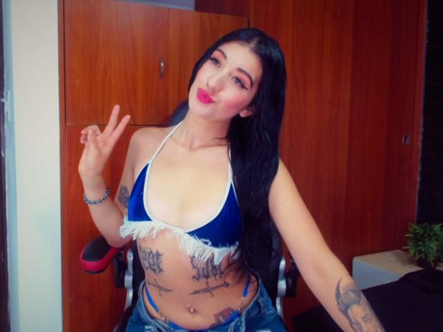 Tifany Diaz, con lunghi capelli neri, indossa una top di velluto blu con frange bianche che scopre il seno, ha tatuaggi sullo stomaco, indossa jeans blu, fa un segno di pace e ha il rossetto rosso..