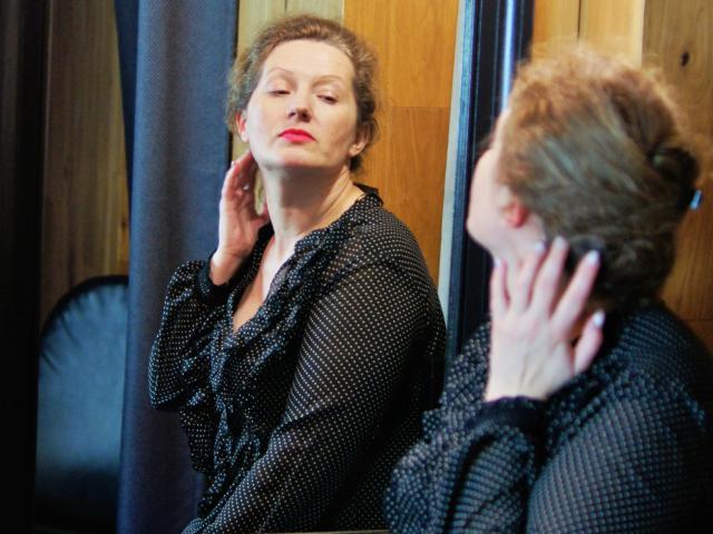 AriaxKiss mit rotem Lippenstift, in einem schwarzen Kleid mit Punkten, steht vor einem Spiegel und berührt ihr Ohr, wobei sich ihr Rücken und ihre Hand darin spiegeln. Ihr Ausdruck ist selbstbewusst..