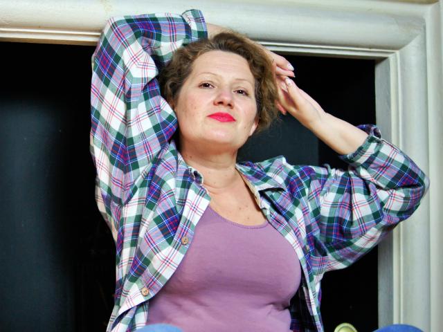 AriaxKiss con cabello castaño rizado, lápiz labial rojo, vistiendo una camiseta morada y una camisa a cuadros azul verdosa, con los brazos levantados, mirando ligeramente hacia un lado, de pie frente a un fondo oscuro..