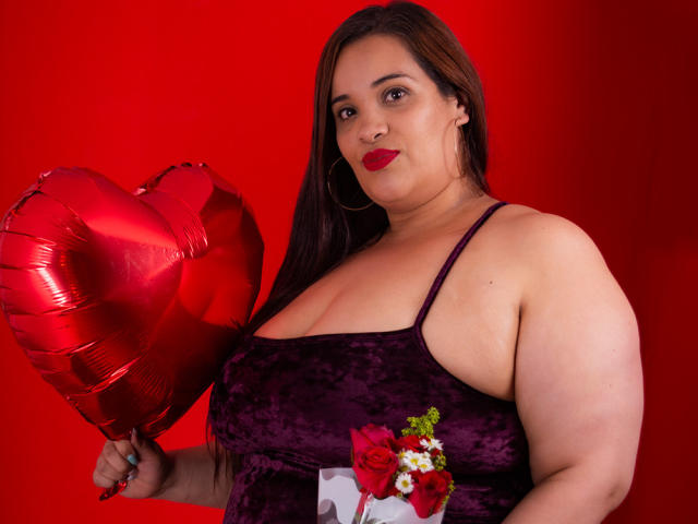 AloondraZaenss with red lipstick, dark brown hair, purple velvet dress, holding shiny red heart balloon, wearing white and red flower on dress, red background, slightly chubby, confident.